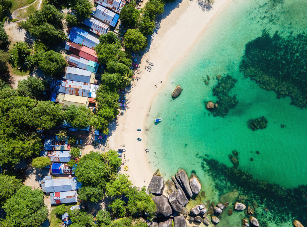 Eine Drohnenaufnahme zeigt einen weißen Sandstrand mit türkisblauem Wasser und kleinen Fischerhütten im Palmenhain.