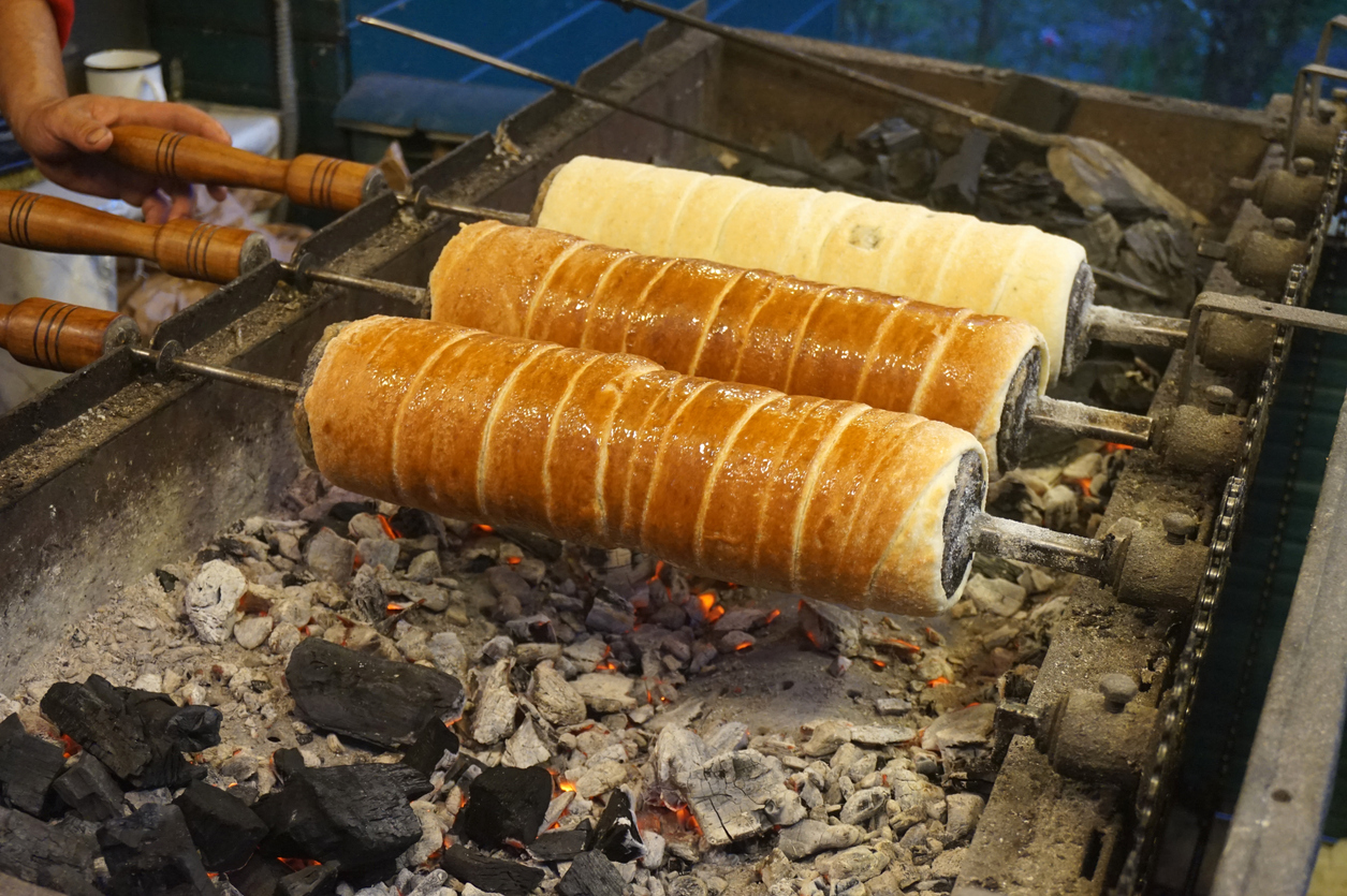 Drei Baumstriezel drehen sich auf Holzrollen über einer offenen Feuerstelle mit glühenden Kohlen.