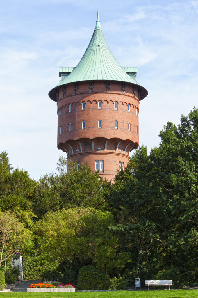 Ein runder Backsteinturm mit grünem, spitzem Dach ragt hinter dichten grünen Bäumen in den blauen Himmel.