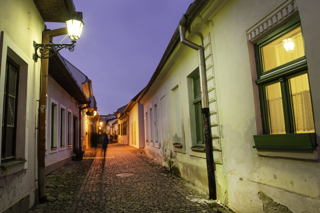 Eine schmale Kopfsteinpflastergasse mit warmen Laternen und Häusern mit Fenstern und Türen bei Dämmerung.