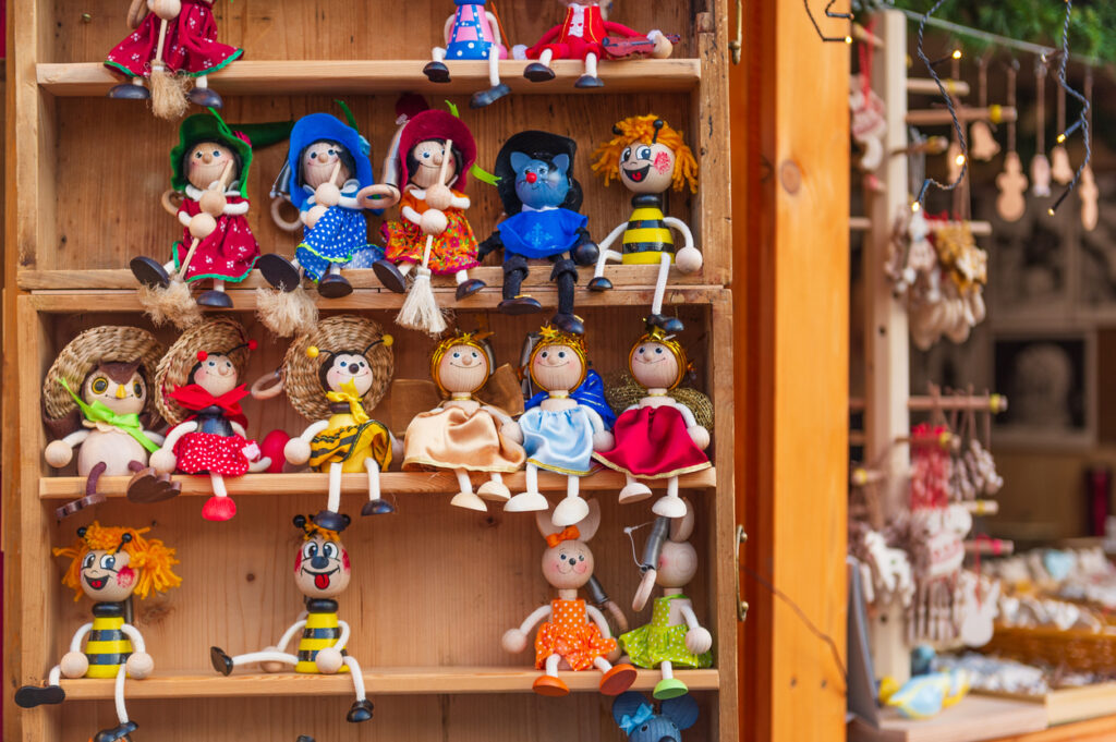Eine Auswahl farbenfroher Holzpuppen und kleiner Figuren sitzt in einem Regal an einem Marktstand.