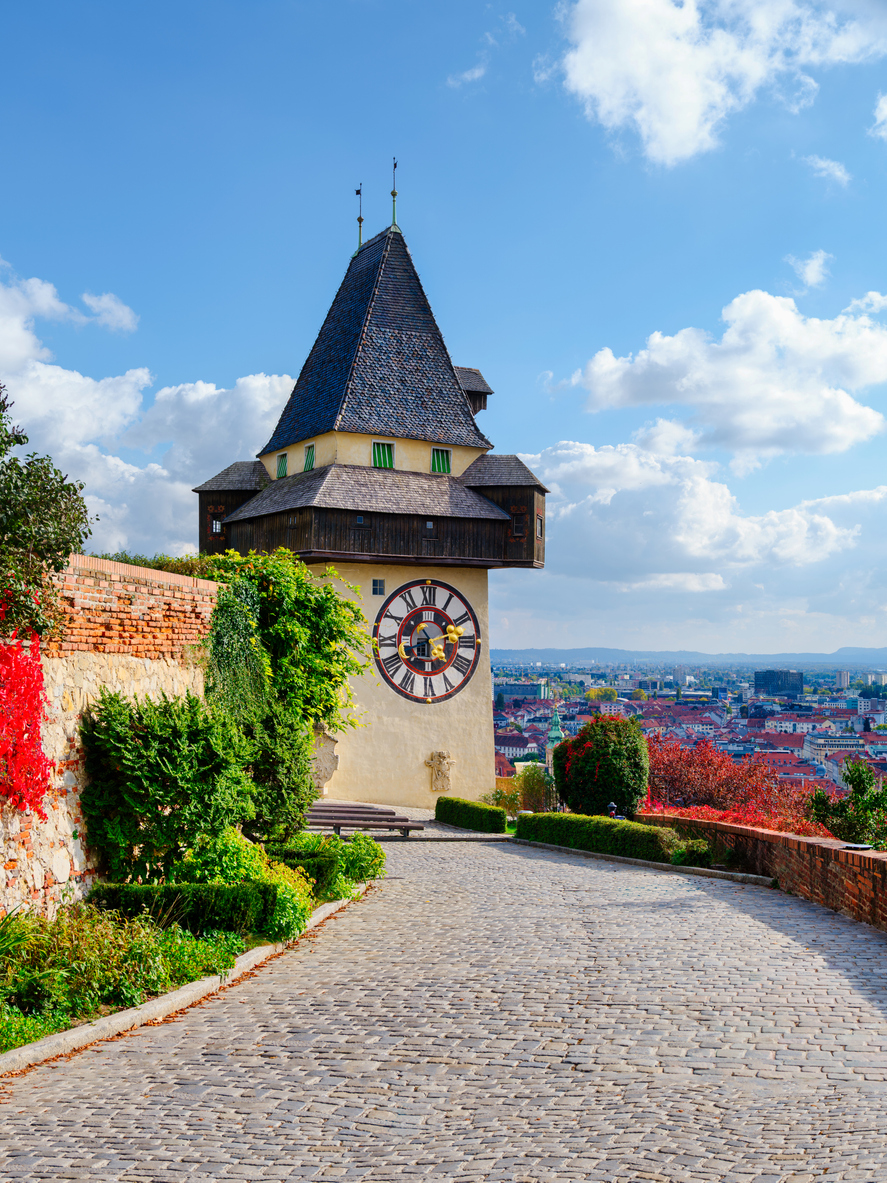 Nahaufnahme des historischen Uhrturms in Graz mit seinem hölzernen Wehrgang vor einem leicht bewölkten Himmel.