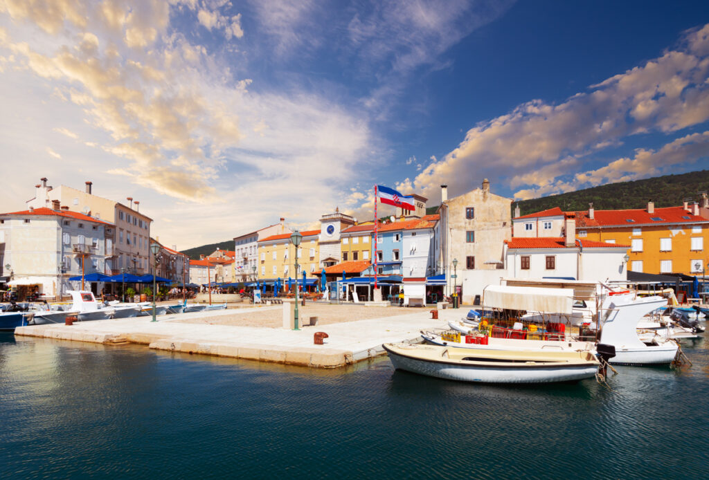 Kleine Fischerboote und Jachten liegen im ruhigen Hafenbecken vor einer Häuserzeile mit dem markanten Stadtturm unter einem pastellfarbenen Himmel.