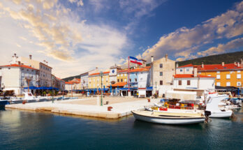 Kleine Fischerboote und Jachten liegen im ruhigen Hafenbecken vor einer Häuserzeile mit dem markanten Stadtturm unter einem pastellfarbenen Himmel.