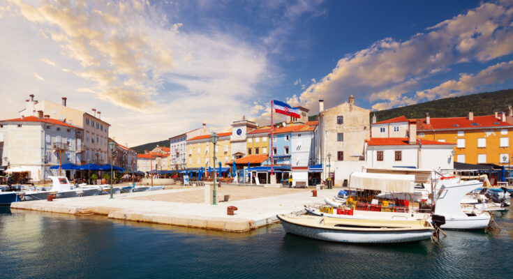 Kleine Fischerboote und Jachten liegen im ruhigen Hafenbecken vor einer Häuserzeile mit dem markanten Stadtturm unter einem pastellfarbenen Himmel.
