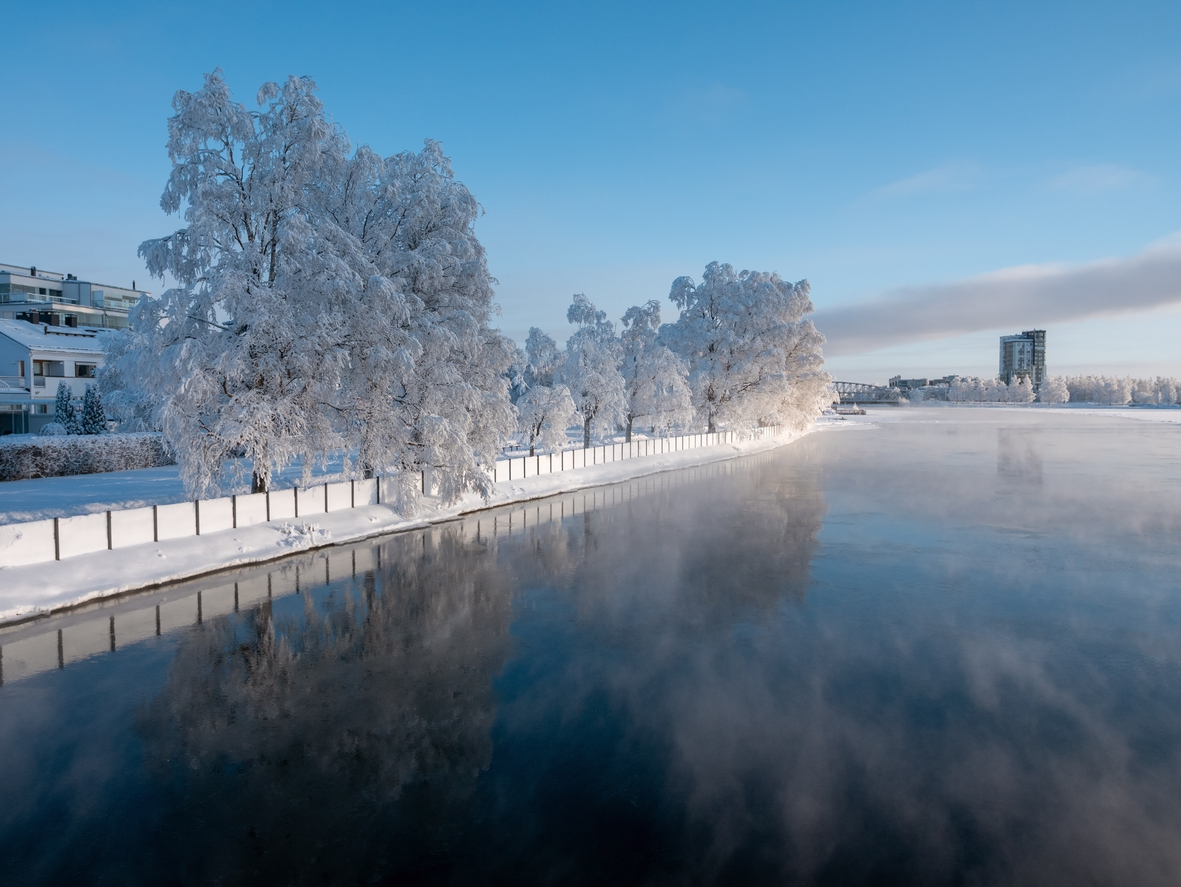 Schneebedeckte Bäume stehen am Ufer eines ruhigen Flusses, dessen dunkle Wasserfläche den Himmel spiegelt.