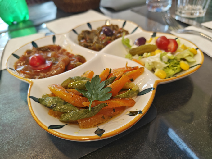 Ein weißer Teller mit vier Fächern präsentiert verschiedene Salate: Karottensticks mit Petersilie, Tomaten-Zwiebel-Mix und Maissalat.