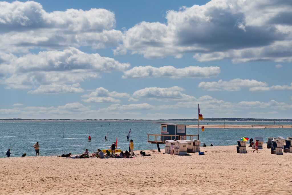 Ein breiter Sandstrand unter blauem Himmel mit weißen Quellwolken; im Vordergrund entspannen Menschen im Sand, während im Hintergrund Strandkörbe und eine Rettungsschwimmerstation zu sehen sind.