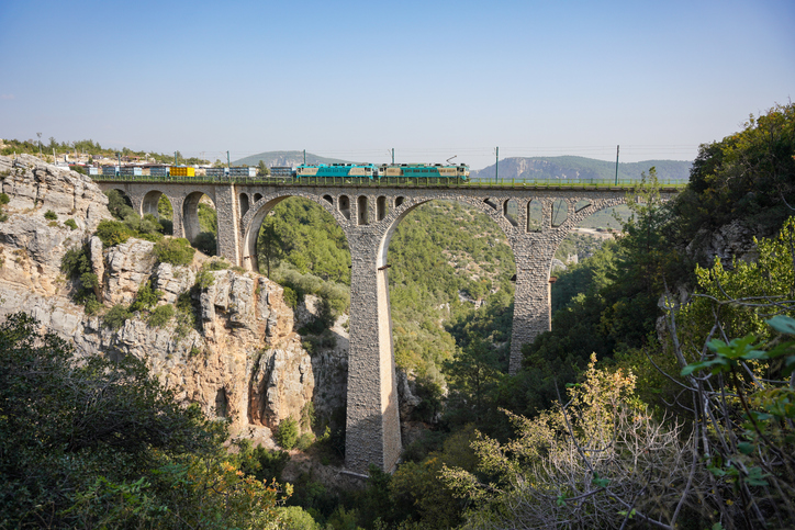 Ein türkisblauer Zug überquert eine gewaltige, aus Stein gemauerte Bogenbrücke in einer tiefen Schlucht.