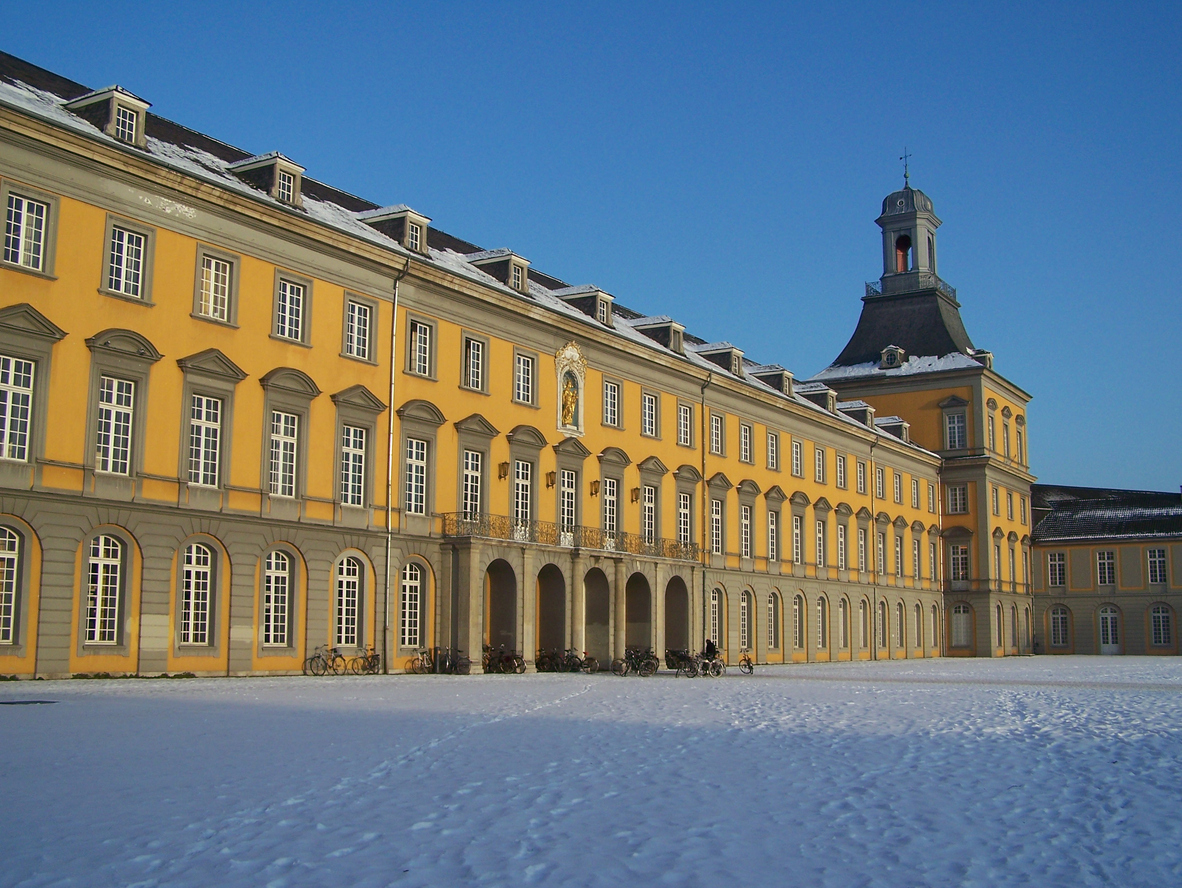 Die gelbe Fassade des Universitätshauptgebäudes erstreckt sich hinter einer weiten, schneebedeckten Fläche unter strahlend blauem Himmel.