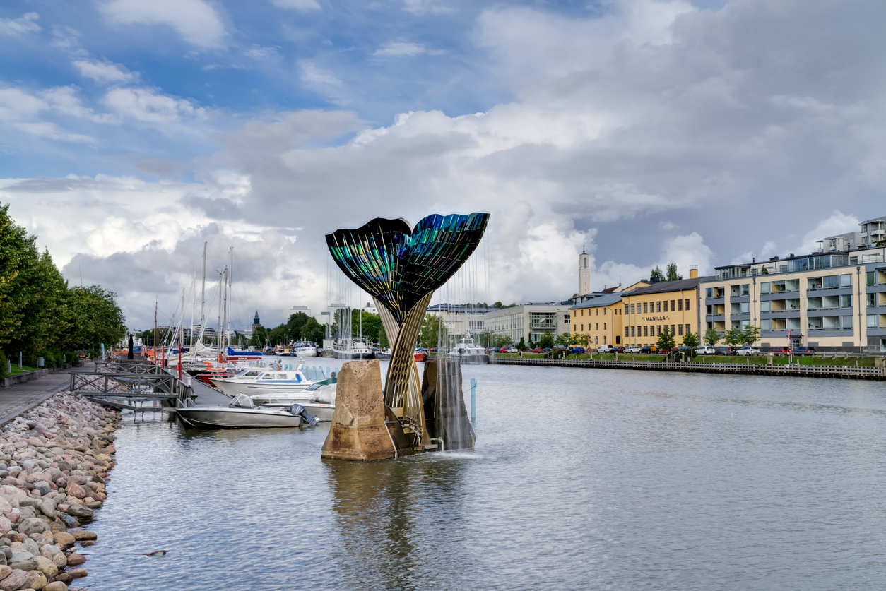 Eine große Metallskulptur in Form einer Walflosse steht direkt im Wasser des Aura-Flusses in Turku, umgeben von Booten und Wohnhäusern.