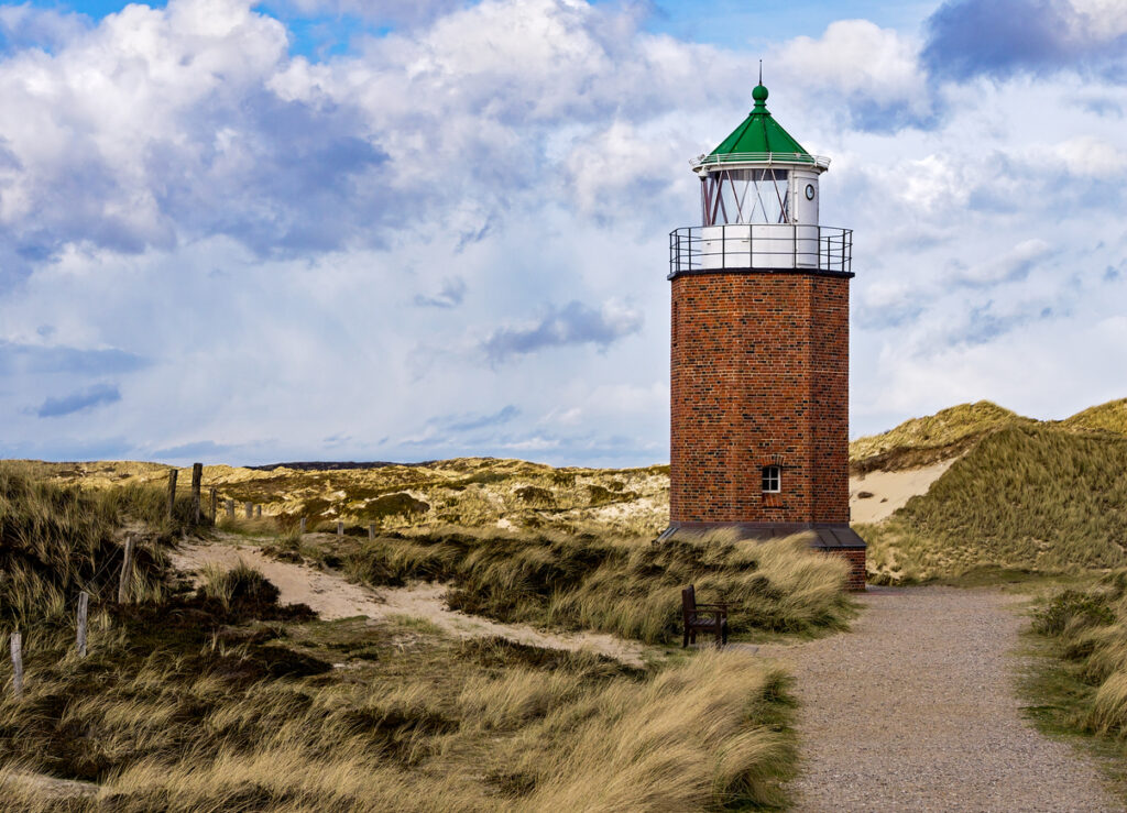 Ein runder Backsteinturm mit grünem Dach steht markant inmitten einer weiten Dünenlandschaft mit trockenem Gras unter einem dramatischen Wolkenhimmel.