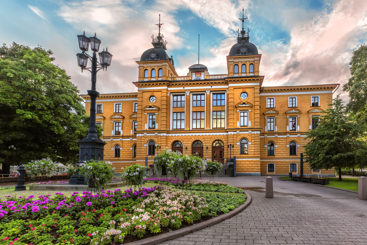 Blick auf ein gelb-orangefarbenes historisches Gebäude mit Türmen, Blumenrabatten und einem Laternenmast im Vordergrund in Oulu.