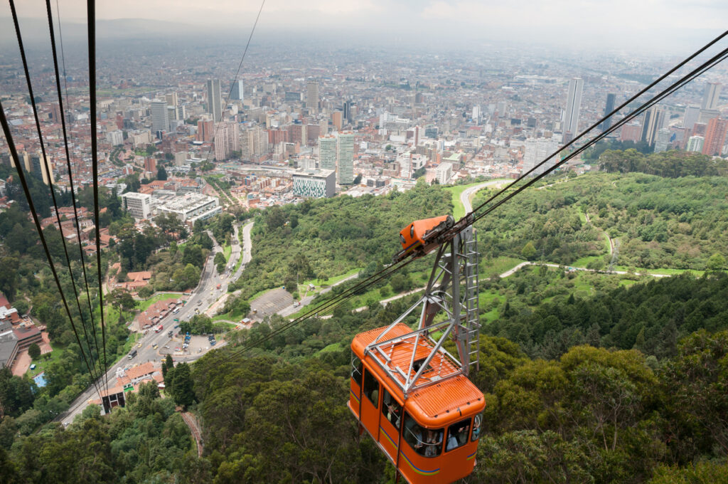 Eine orangefarbene Seilbahnkabine schwebt über dichten grünen Bäumen mit Blick auf die riesige Stadt Bogotá.