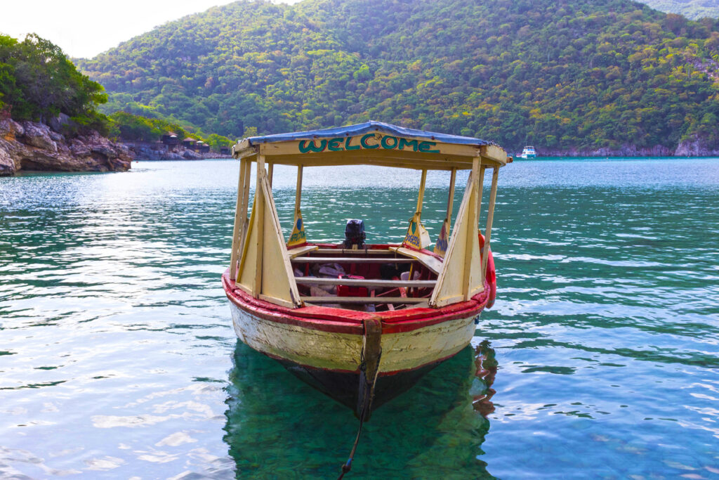 Ein buntes Holzboot mit der Aufschrift „WELCOME“ ankert im türkisblauen Wasser vor bewaldeten Hügeln.