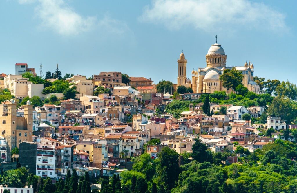 Blick über dicht bebaute Hügel in Algier mit auffälliger Basilika und grünen Hängen.