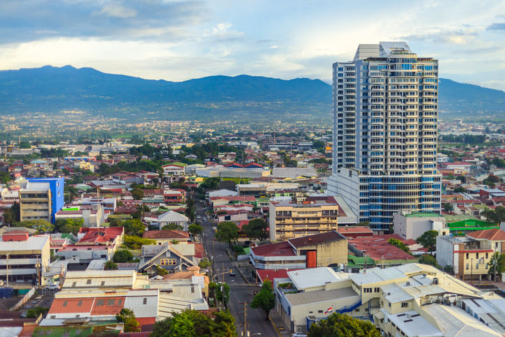 Ein Blick über die Dächer von San José mit einem markanten modernen Hochhaus im Vordergrund und den grünen Bergen des Zentraltals im Hintergrund.