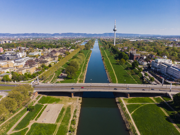 Eine Luftaufnahme zeigt den geradlinigen Verlauf des Neckars durch Mannheim, flankiert von grünen Ufern, einer Brücke im Vordergrund und dem Fernmeldeturm im Hintergrund.
