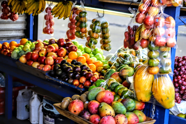 Ein bunter Marktstand präsentiert eine Fülle an tropischen Früchten wie Bananen, Mangos, Papayas und Äpfeln in Körben und Beuteln.