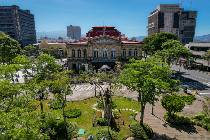 Blick von oben auf das Teatro Nacional de Costa Rica mit seinem roten Dach, umgeben von grünen Bäumen und der Plaza de la Cultura.