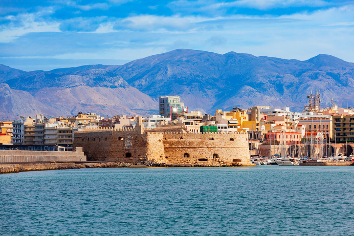 Eine massive mittelalterliche Steinfestung am Ende einer Kaimauer im Meer, dahinter die Skyline von Heraklion vor einer Gebirgskette.
