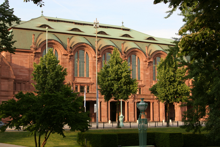 Ein imposantes historisches Gebäude aus rotem Sandstein mit einem hellgrünen Kupferdach und großen Rundbogenfenstern vor grünen Bäumen.