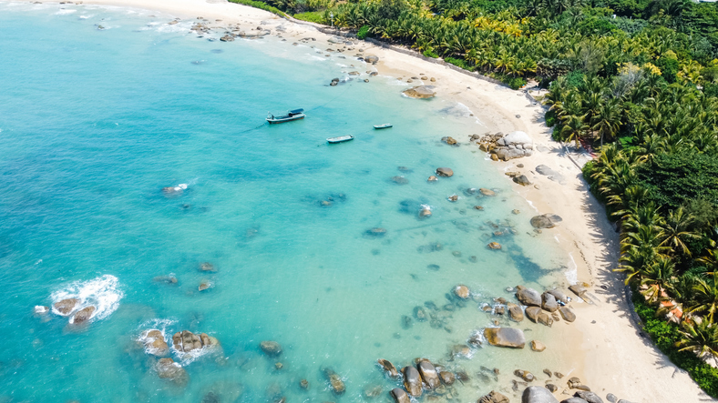 Eine Luftaufnahme zeigt große Granitfelsen im flachen, glasklaren Wasser eines Sandstrandes, der von dichten Palmenwäldern gesäumt wird.
