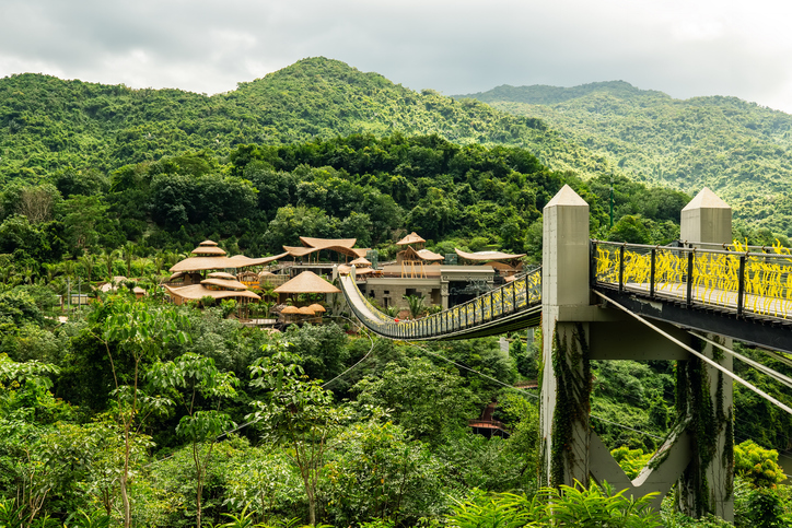 Eine lange, schmale Hängebrücke spannt sich über ein dichtes, grünes Dschungeltal hinweg auf bewaldete Berge zu.