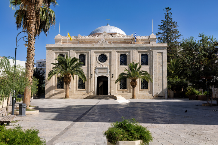 Die helle Fassade der Kirche Agios Titos an einem sonnigen Platz, flankiert von zwei Palmen unter einem wolkenlosen blauen Himmel.