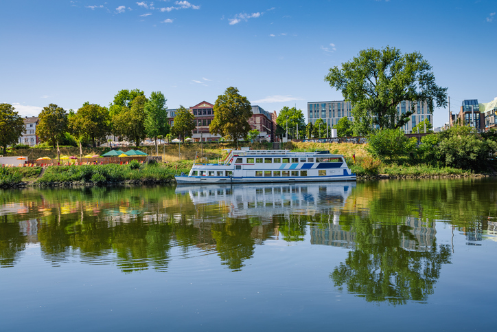Ein weiß-blaues Fahrgastschiff fährt auf dem Neckar an einem Stadtstrand mit bunten Sonnenschirmen und grünen Bäumen vorbei.