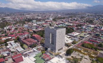 Eine Luftaufnahme zeigt ein markantes, graues Betongebäude mit einer großen rechteckigen Öffnung in der Mitte, umgeben von der weitläufigen Stadtbebauung.