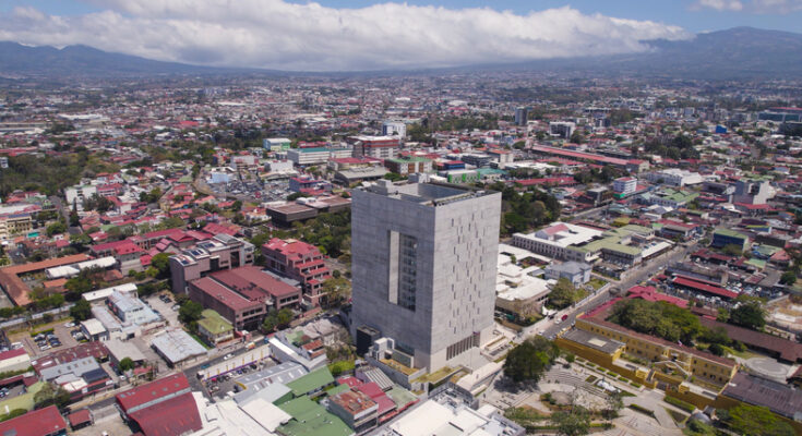 Eine Luftaufnahme zeigt ein markantes, graues Betongebäude mit einer großen rechteckigen Öffnung in der Mitte, umgeben von der weitläufigen Stadtbebauung.