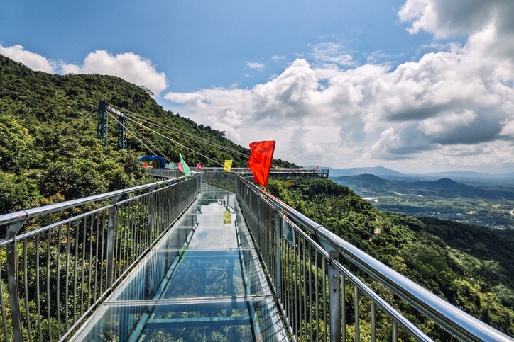 Ein gläserner Skywalk führt entlang einer bewaldeten Bergflanke; im Hintergrund öffnet sich ein weiter Blick über das grüne Tal unter dramatischen Wolken.