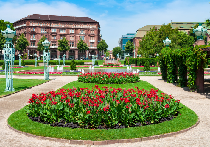 Ein kreisrundes Beet mit leuchtend roten Blumen im Vordergrund einer gepflegten Parkanlage mit historischen Arkadengebäuden und Wasserfontänen.
